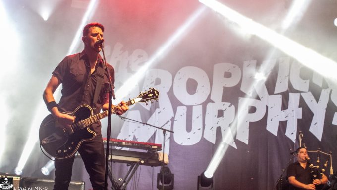 A musician plays guitar on stage with bright lights and a band backdrop.