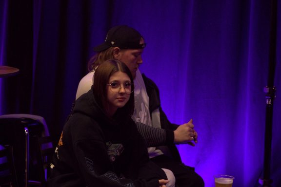 Two people seated on stage, one in a baseball cap and the other in a hoodie, with purple lighting.