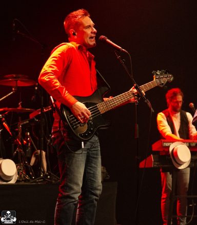 Man singing on stage with a guitar, accompanied by a musician at a keyboard.