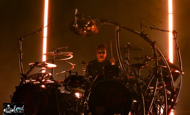 Drummer performing on stage, surrounded by illuminated instruments and a vibrant atmosphere.
