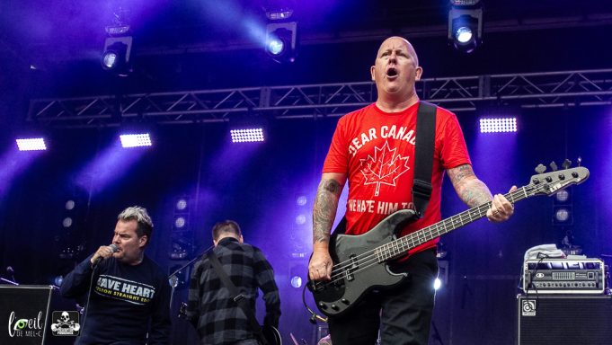 Two musicians perform energetically on stage with bright stage lights in the background.