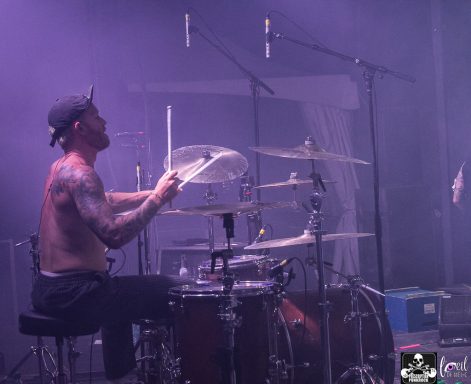 A drummer performing on stage with a purple-lit background.