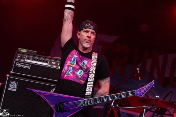 A musician wearing a black headband and a pink T-shirt plays a purple guitar on stage.
