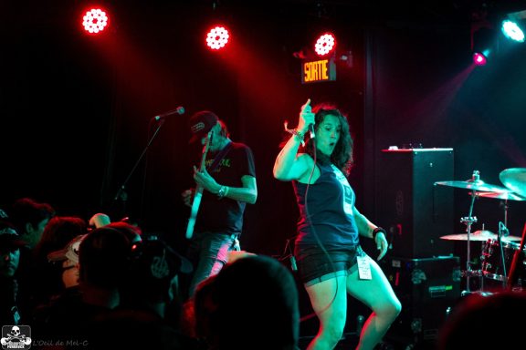 A woman performs energetically on stage with a guitarist; vibrant stage lights create a lively scene.