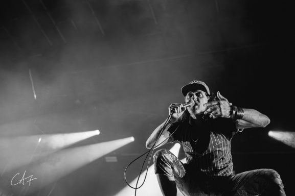 A performer energetically singing on stage with dramatic lighting behind.