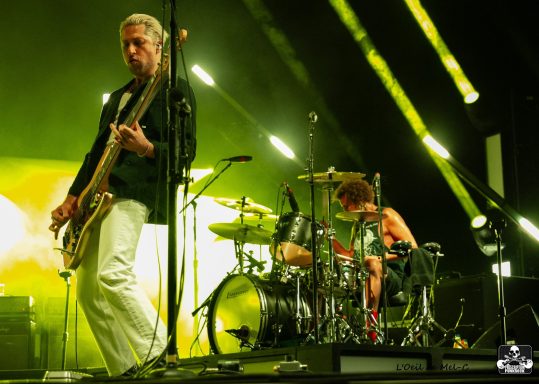 Band performing on stage with a bassist and drummer under vibrant lighting.