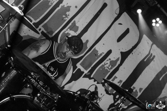 Drummer performing on stage, focused, with a large band name background.