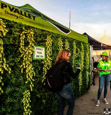A woman stands near a green wall decorated with hops at an outdoor event.