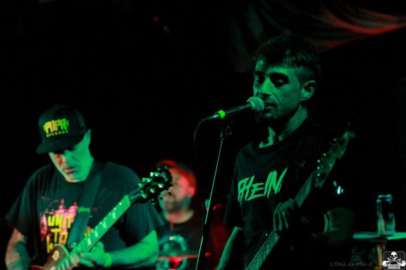 Band performing on stage, with musicians playing guitar and singing under colored lights.