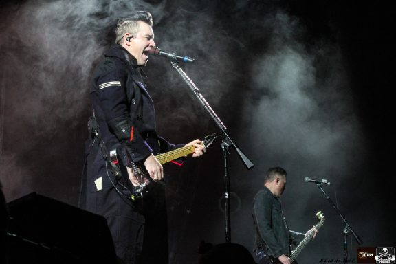 Two musicians perform on stage, with one singing and playing guitar amid smoke.