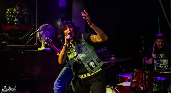 Lead singer in a denim vest performs energetically on stage with a drummer behind.