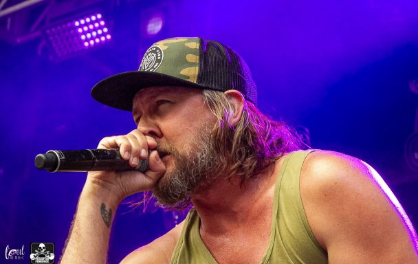 Male singer with long hair, wearing a cap, performs passionately on stage.