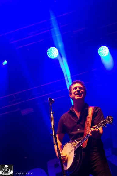 Man smiling and playing a banjo on stage with colorful lighting in the background.