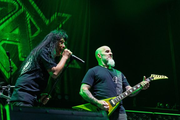 Two male musicians perform on stage with a microphone and guitar, illuminated by green lights.