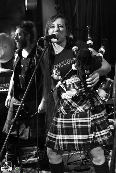 A musician in a kilt plays the bagpipes while singing on stage.