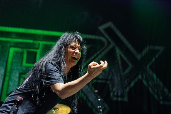 A performer energetically gestures while singing on stage, with a vibrant backdrop.