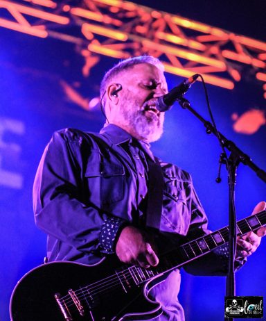 Musician performing on stage with a guitar and microphone, vibrant lighting in the background.