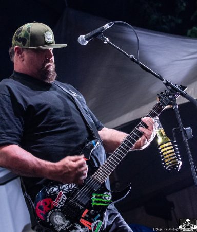 Musician playing electric guitar on stage, wearing a cap and a black shirt.