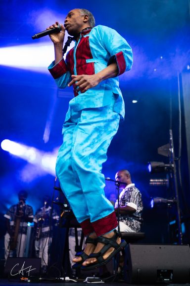 A performer in a blue outfit jumps while holding a microphone on stage.