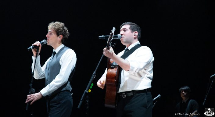 Two performers on stage, one singing into a microphone and the other playing guitar.