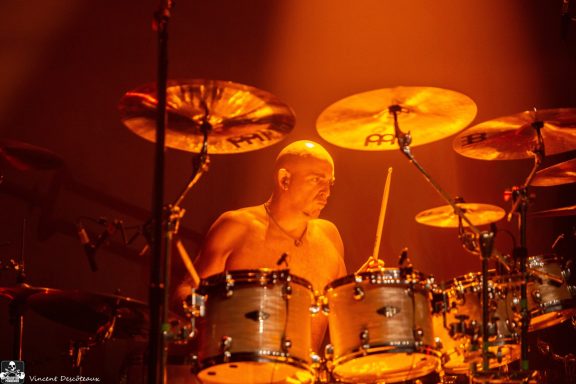 A drummer performs energetically under warm stage lighting.