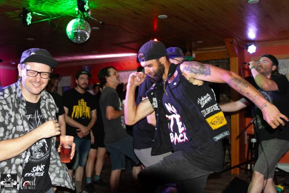 A lively crowd at a music event, with a man dancing and another smiling, holding a drink.