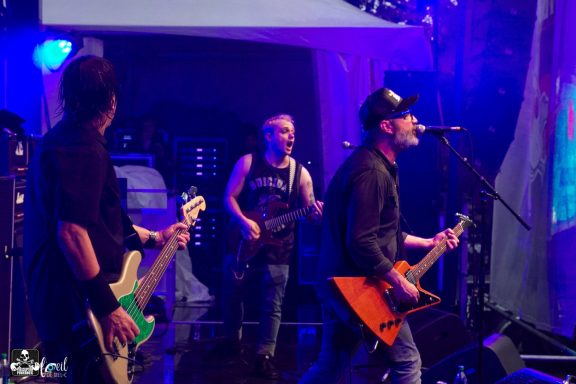 Three musicians performing on stage with instruments, under colorful stage lighting.