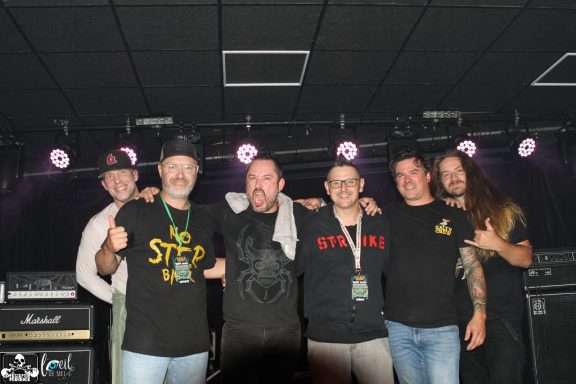A group of six men posing together on stage with musical equipment in the background.