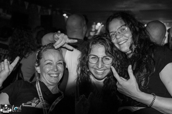 Three smiling women pose together, making hand gestures in a lively atmosphere.