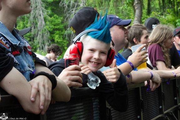 Child with blue mohawk hairstyle and ear protection giving thumbs up at a crowded event.