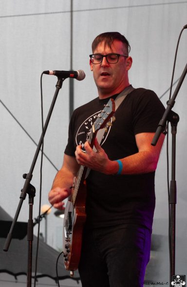 Musician performing on stage with a guitar and microphone, wearing glasses and a black t-shirt.