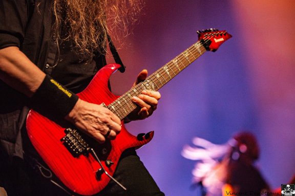 A musician playing a red electric guitar on stage, with dramatic lighting in the background.