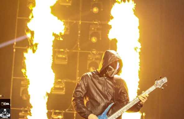 Musician in a hooded costume playing a blue bass guitar with flames in the background.