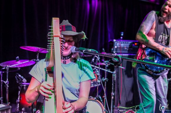 Musician playing a harp with a guitarist in the background, performing on stage.
