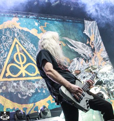 A musician with long white hair plays guitar in front of a vibrant backdrop.