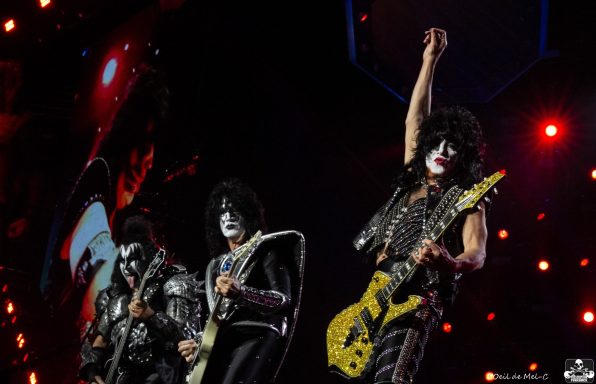 Three musicians in rock attire perform on stage, one raising a guitar triumphantly.
