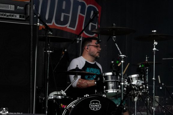 Drummer performing on stage with a drum set, wearing glasses and a black and white shirt.