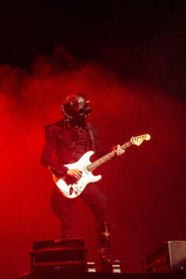 Musician in a black outfit and mask playing an electric guitar against a red backdrop.
