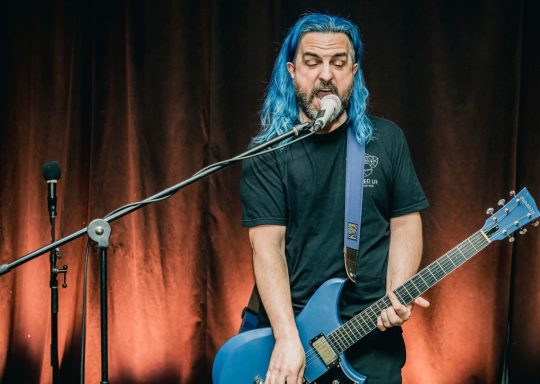Rouge Pompier - Radiothon CKRL 89,1 Musician with blue hair performing on stage, playing a blue electric guitar.
