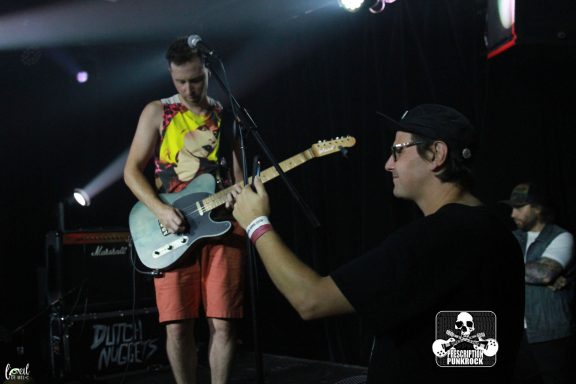 Musician playing guitar on stage with another person taking a photo.