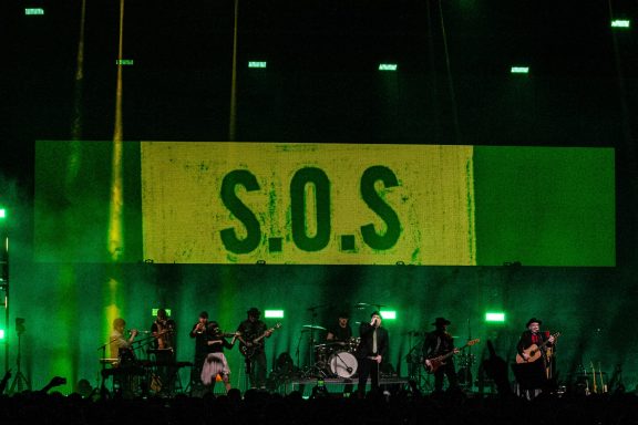 A live concert scene with performers on stage and a large "S.O.S" sign behind them.