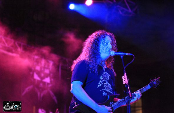 A musician with curly hair performs on stage with a guitar, illuminated by colorful lights.