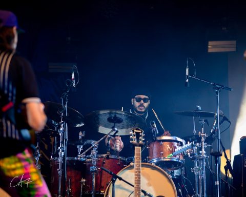 Drummer performing on stage with colorful band members in the background.