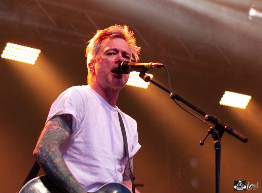 A musician with tattoos sings into a microphone on stage, with bright lights in the background.