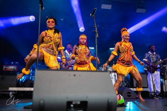 Three dancers in vibrant costumes performing on stage with a band in the background.