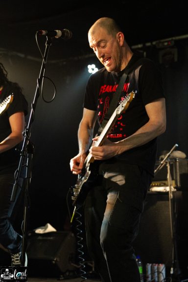 Dead Bob - La Source de la Martinière Musician playing guitar on stage, wearing a black T-shirt, with intense expression.