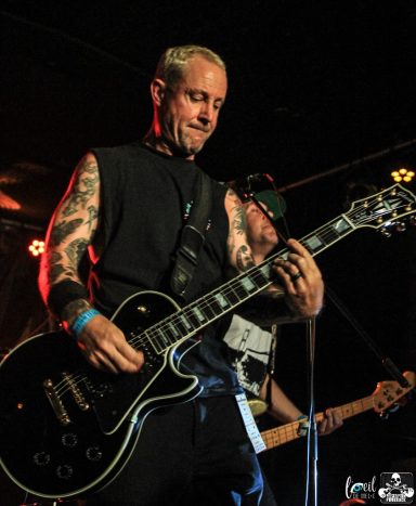 Man playing an electric guitar on stage with tattoos and a focused expression.