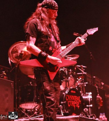 A guitarist with long hair playing a red electric guitar on stage.