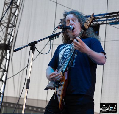 Musician with curly hair performs on stage, playing a guitar and singing.