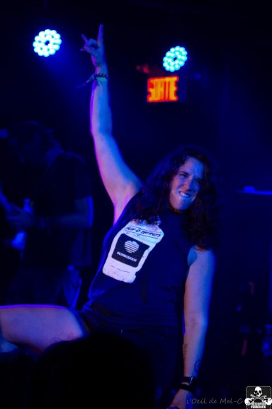 Person with dark hair poses confidently on stage while holding one arm up, illuminated by blue lights.